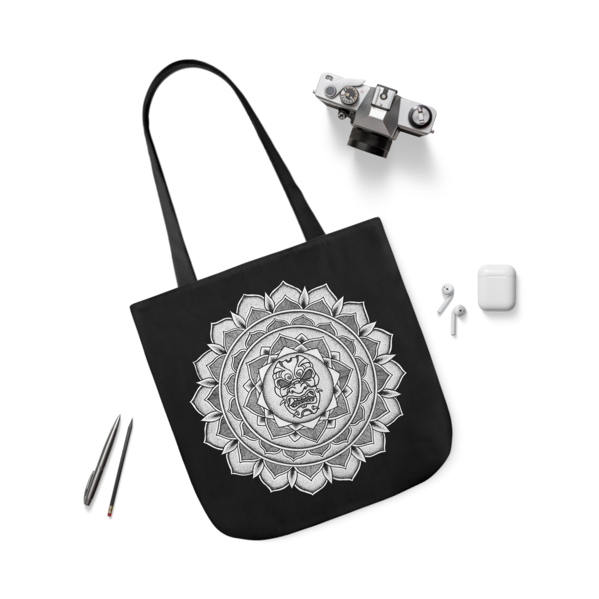 image of a black mandalarian brand tote bag with black handle and a white and black samurai mask design print on both sides next to a camera and earphones and a pen and pencil