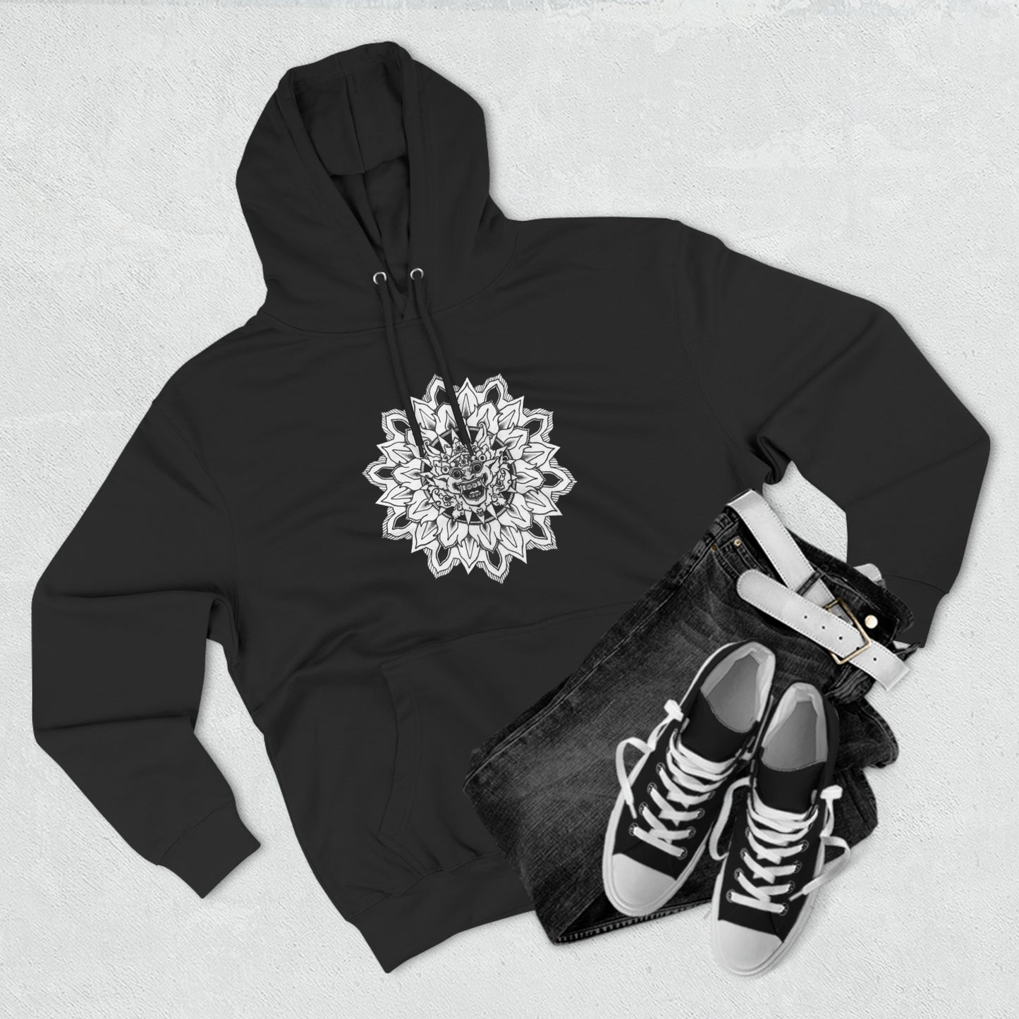 image of a unisex black hooded sweatshirt with white and back bhoma bali god mandala design on the front next to some jeans and sneakers