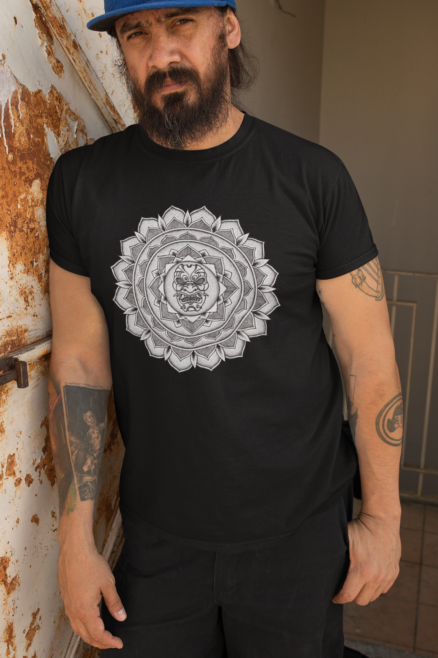 a bearded man leaning against a rusty wall wearing a black mandalarian brand tshirt with samurai mask mandala tattoo design 