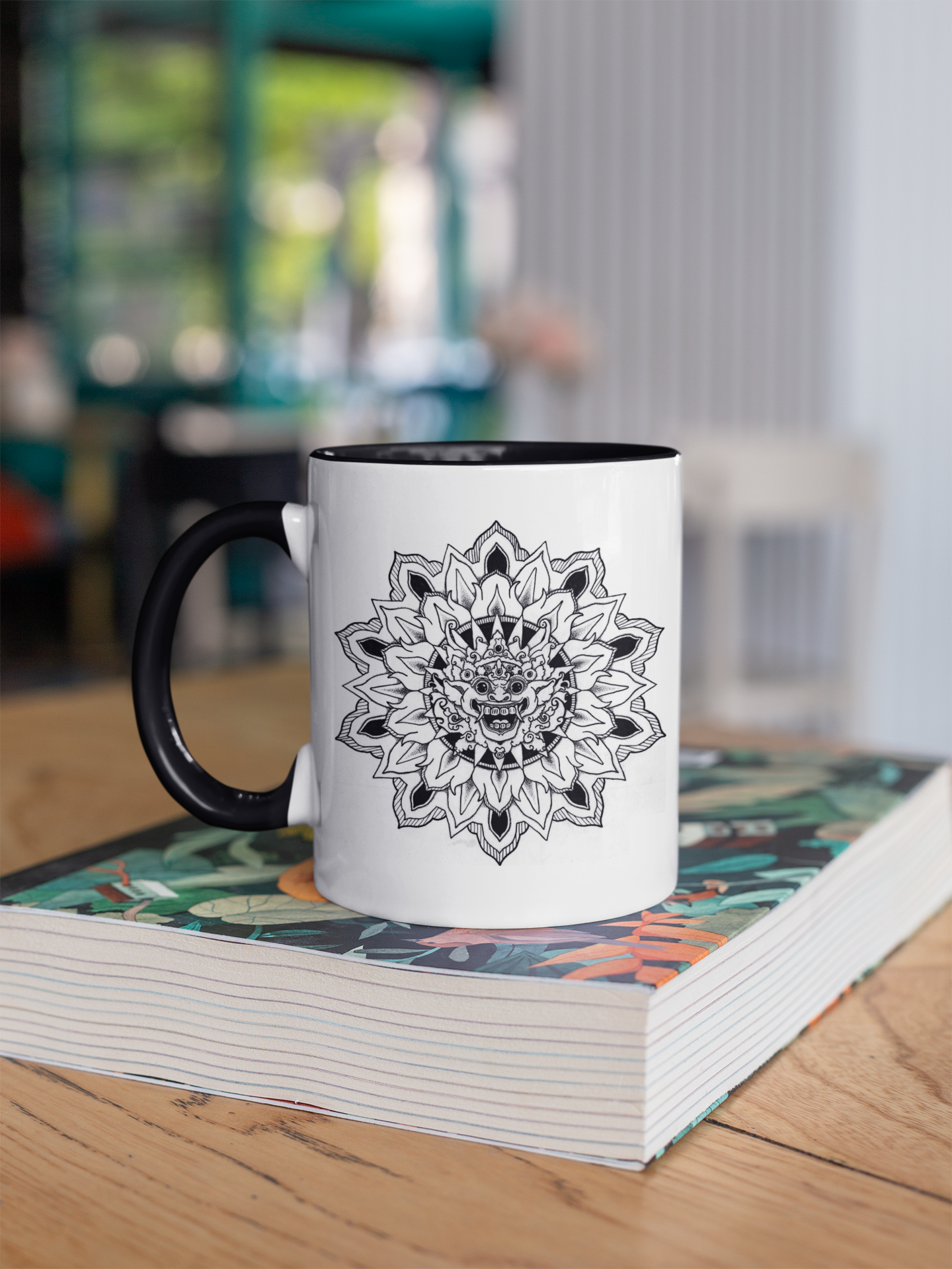 a two toned white and black 11oz coffee mug with bhoma bali god mandala design resting on a book on a table