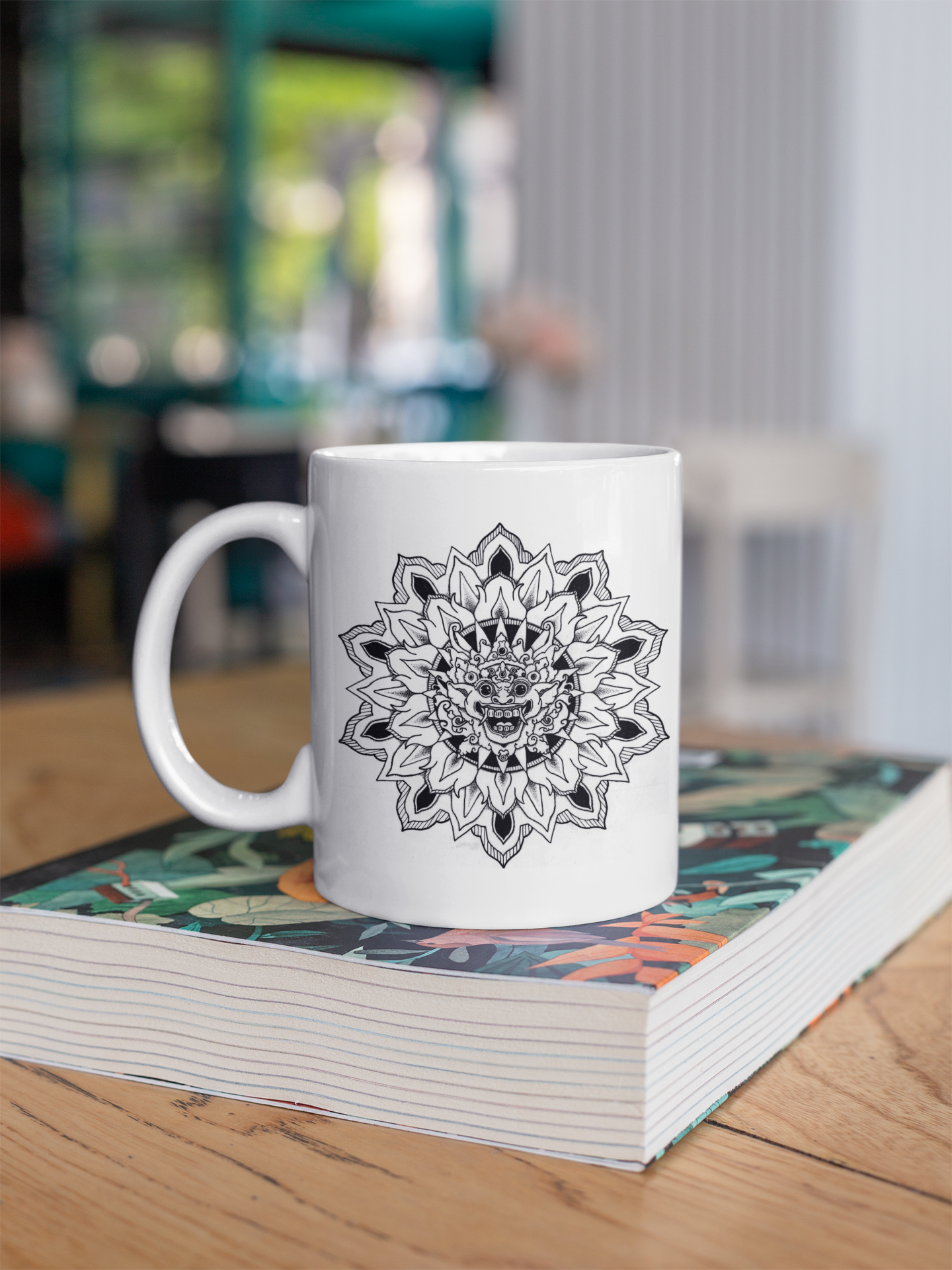 a white 11oz coffee mug with black bhoma bali god mandala design resting on a book on a coffee table