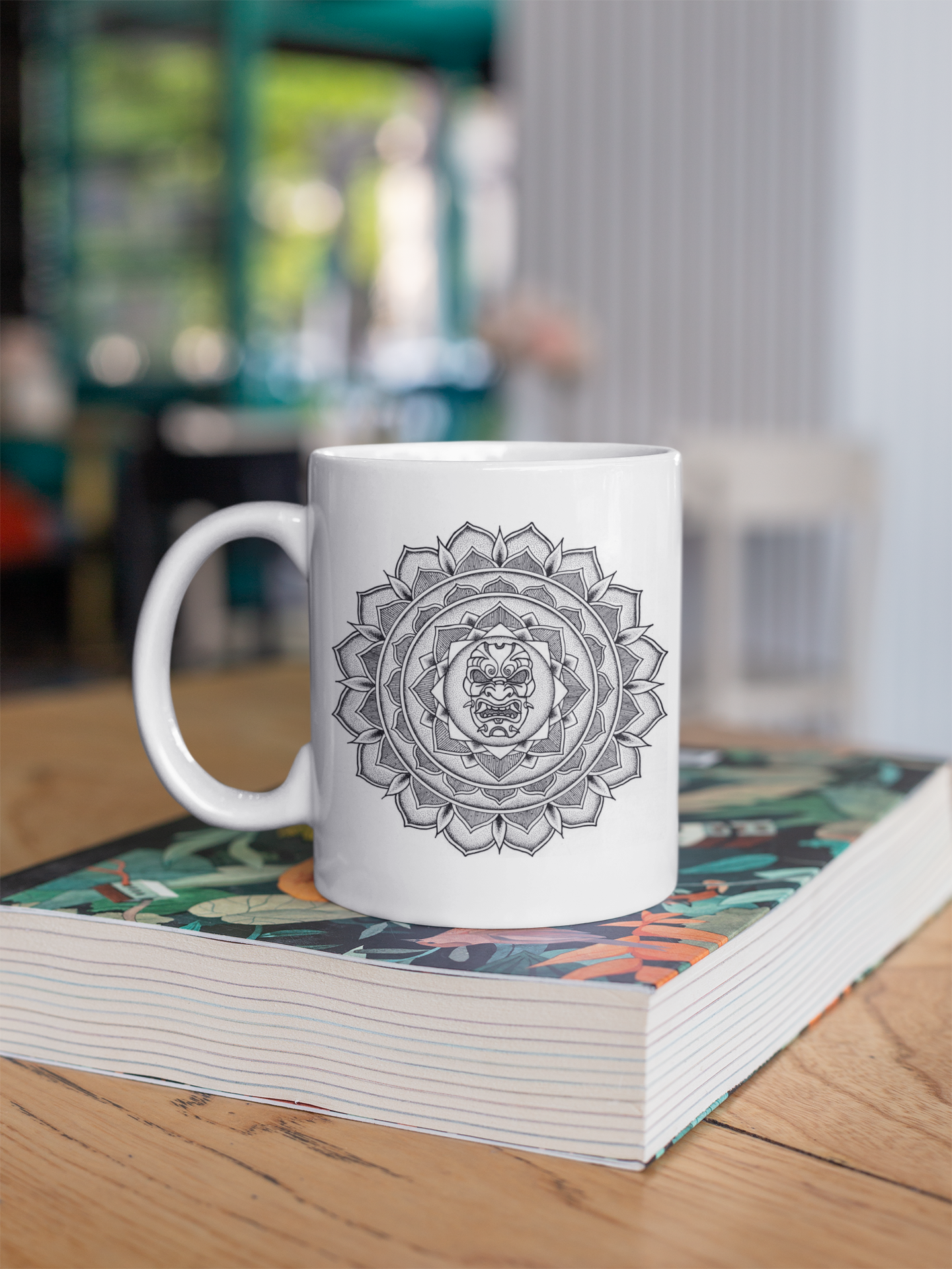 a white 11oz coffee mug with black samurai mask mandala design resting on a book on coffee table