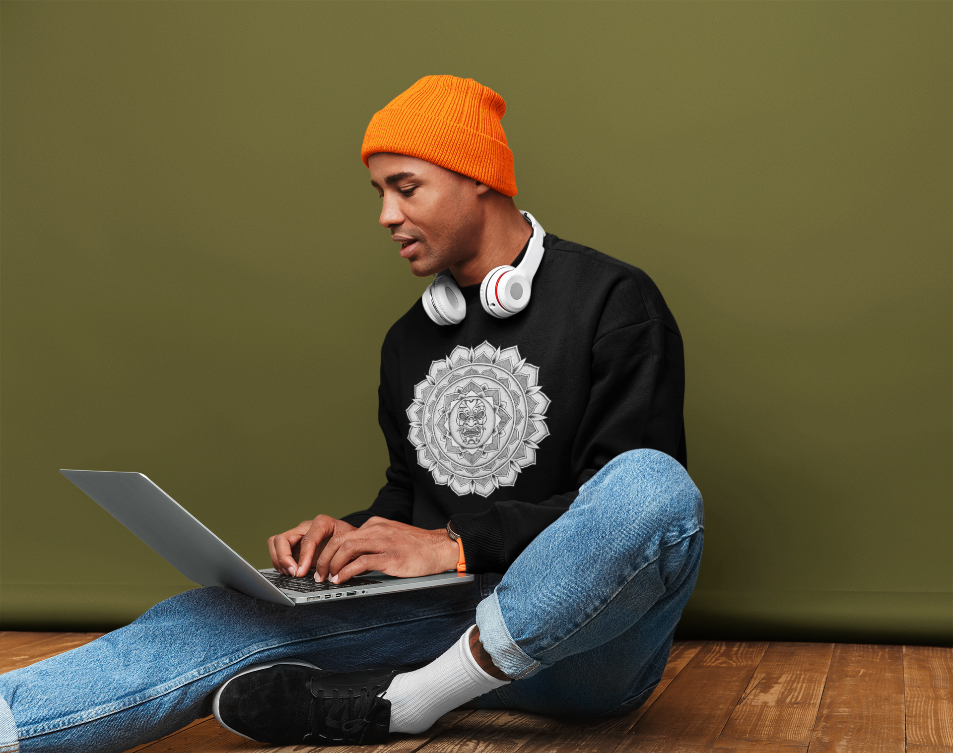 image of a man sat on the floor inside a house working on a laptop wearing an orange beanie and blue jeans and a black mandalarian brand sweatshirt with a white and black samurai mask mandala design on the front