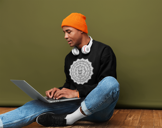 image of a man sat on the floor inside a house working on a laptop wearing an orange beanie and blue jeans and a black mandalarian brand sweatshirt with a white and black samurai mask mandala design on the front