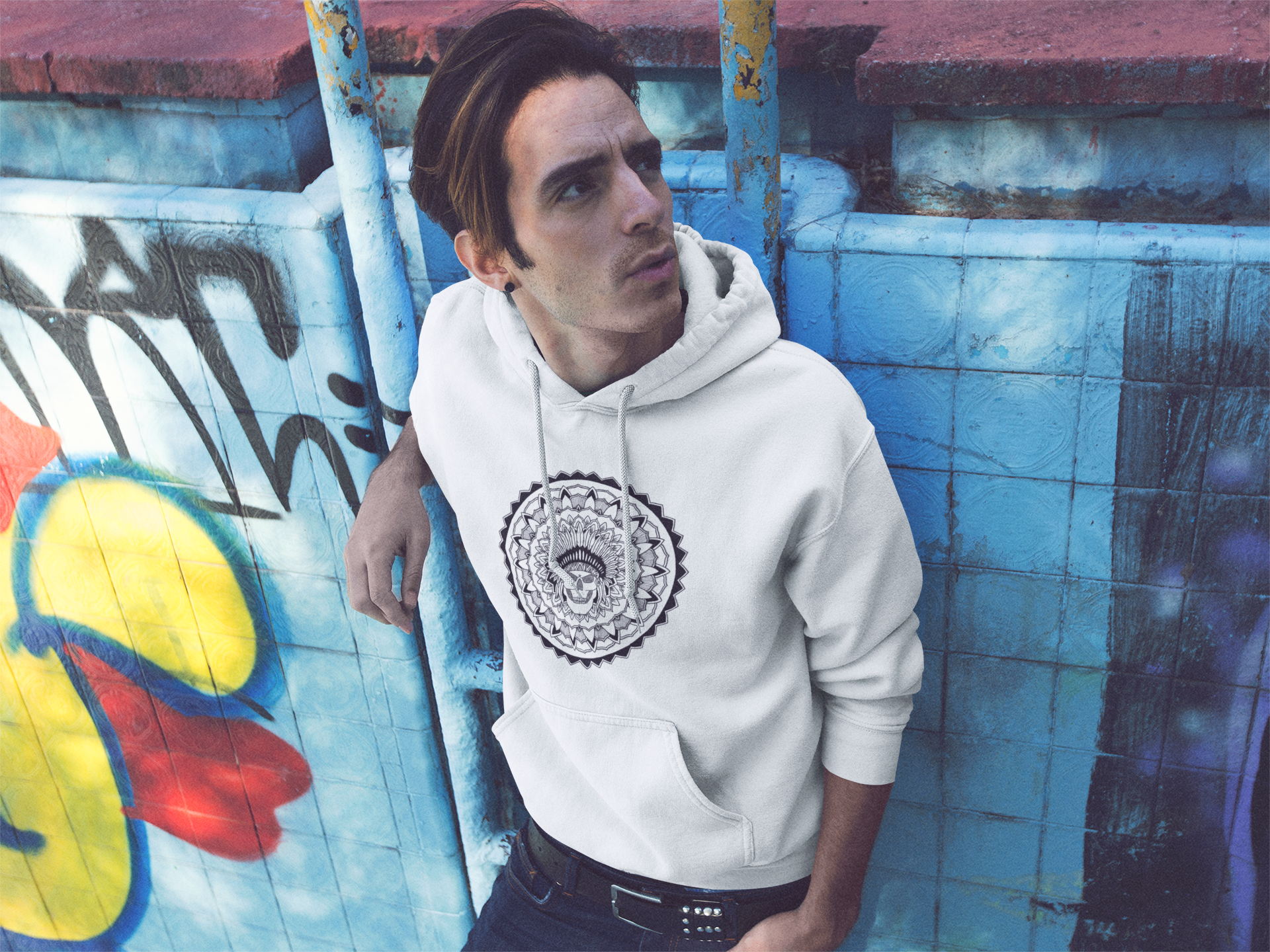 trendy guy leaning against graffiti wall wearing a white mandalarian brand hoodie with chieftain skull design